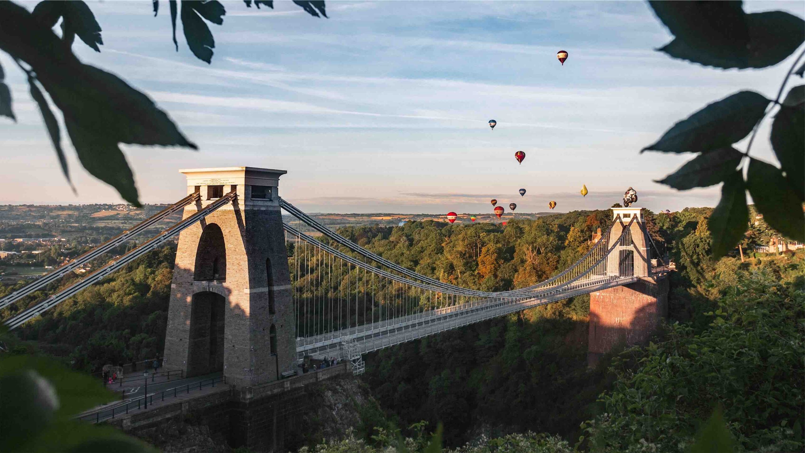 Clifton suspension bridge at sunset with air balloons in the sky above.