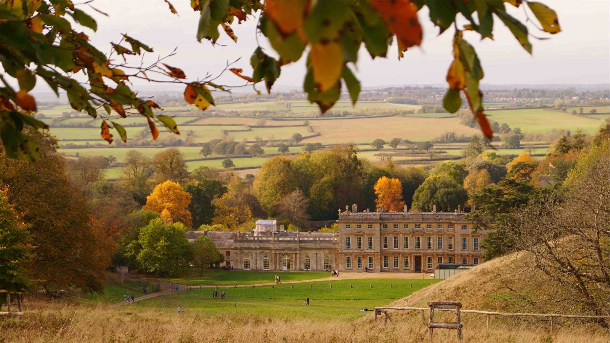 Grand estate house sits down a hill with golden trees and fields surrounding the grounds