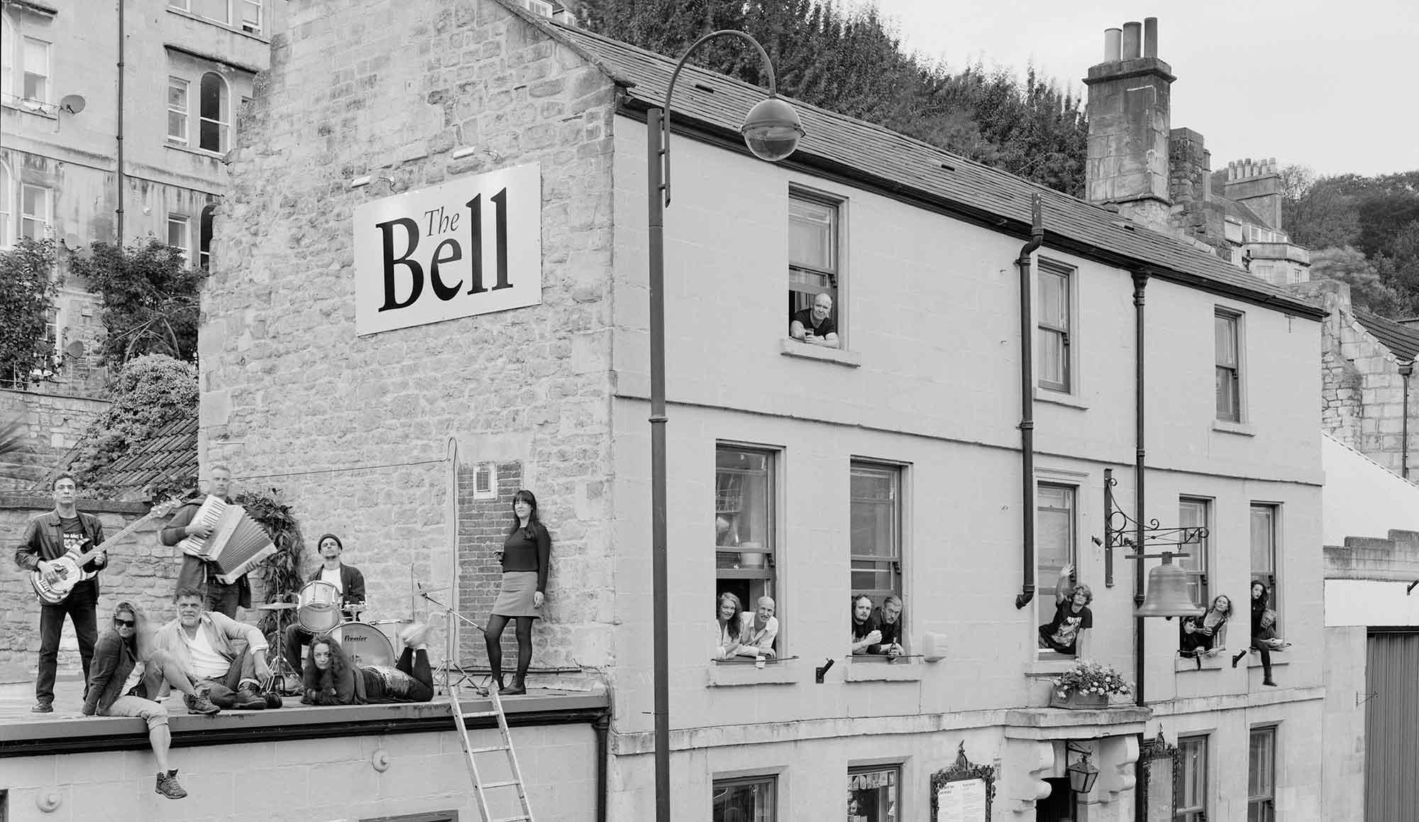 The Bell pub Bath showing the side and front of the building. People are poking through the windows who appear to work at the pub. People on the roof next door appear to be a band with musical instruments.