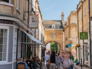 The Raven pub bath sign and facade. The street begins to get narrower and a large arch with window above. People walk down the street and sitting on chairs outside of the pub.