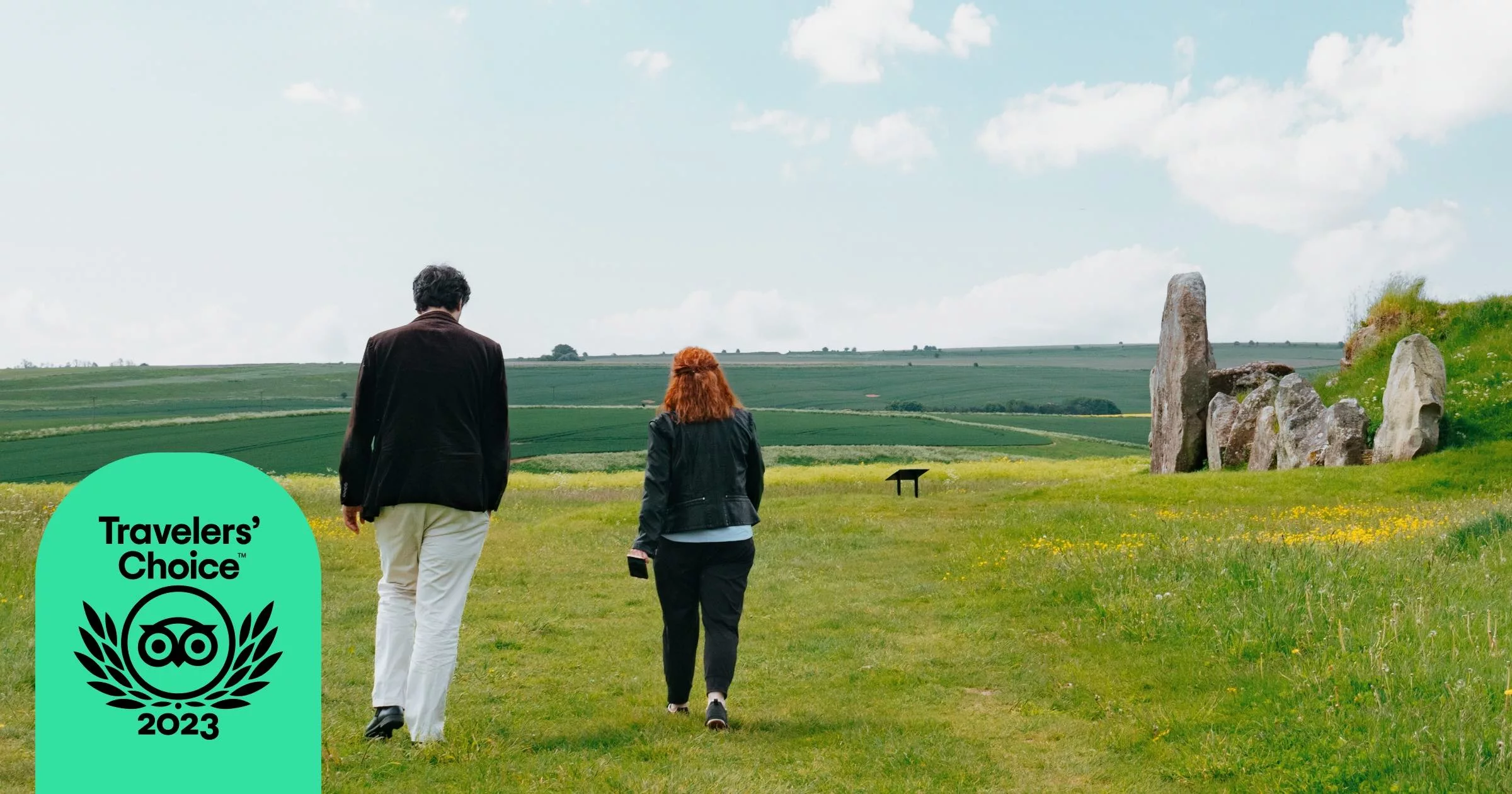 2 people in frame with their backs turned as they walk towards stone formation. Grass fields surround the two subjects.