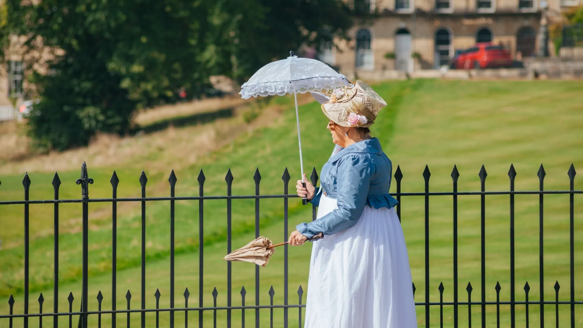Two women in period clothing dress from the Jane Austen era. We see them from their side profiles in front of railings with green grass behind.