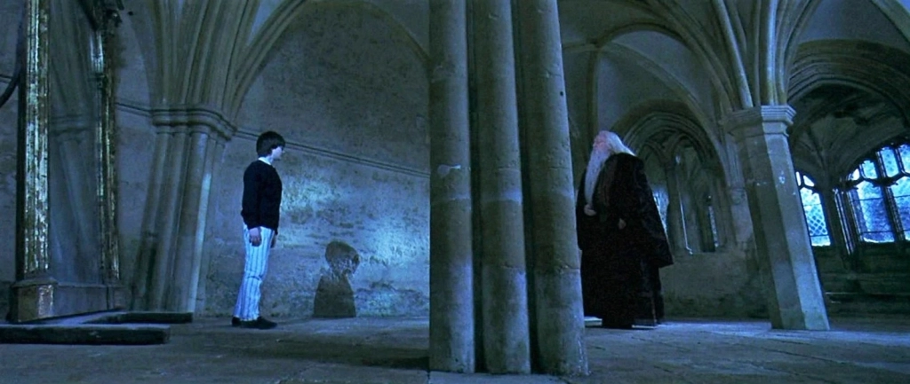 A film scene from Harry Potter and the Philosopher's Stone when Harry encounters the mirror or Erised. The room is old and empty with large archways. To the left stands a tall mirror with Harry in front facing Dumbledore who stands to the right of the frame.