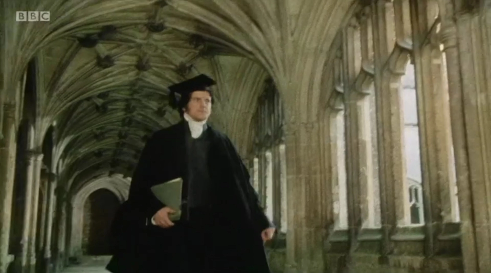 Colin Firth as Mark Darcy in Oxford dress robes in a grand hallway with large arches.