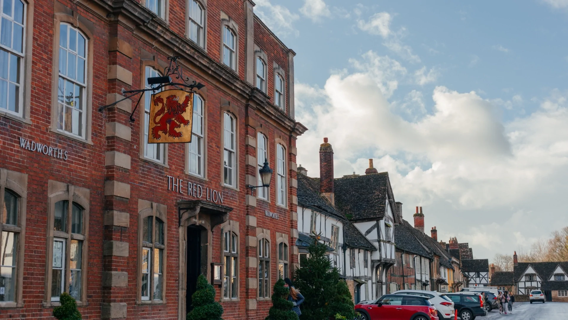 The Red Lion pub is positioned on the left-hand side of the frame where a gold sign with a red lion on the facade. On the right a street with cars and old houses till the end of the frame.