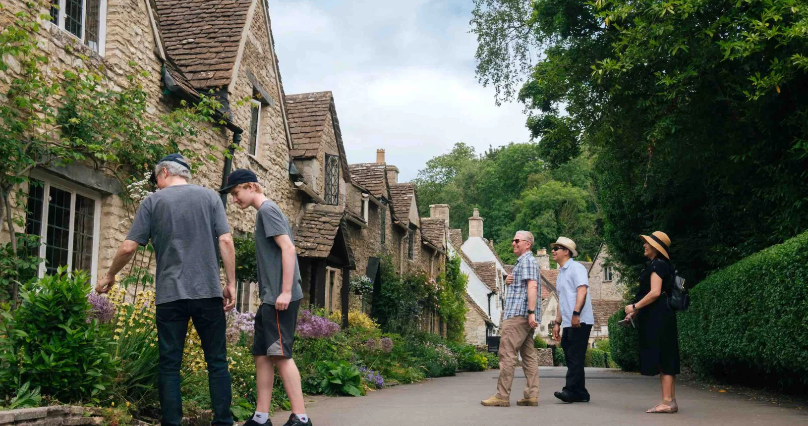 Quaint house line the street on the left side. A father and son stand near the Cotswold houses and a group of three people in the background smiling.