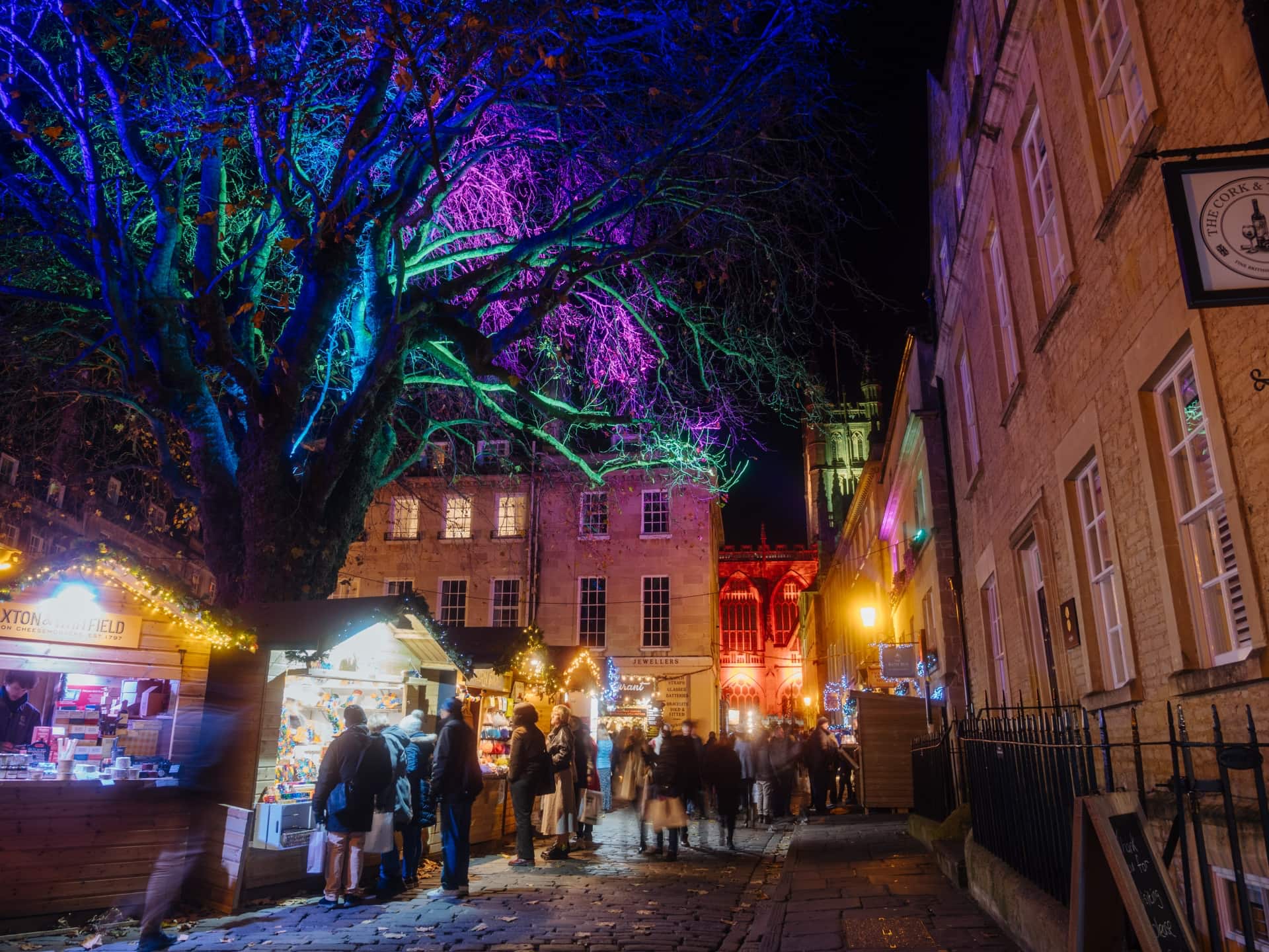 Wooden huts selling Christmas gifts to people on a cobbled street. It is night and a large tree behind the stalls is lit up in multicolour. Classic Bath stone houses line the street as people wiz by.