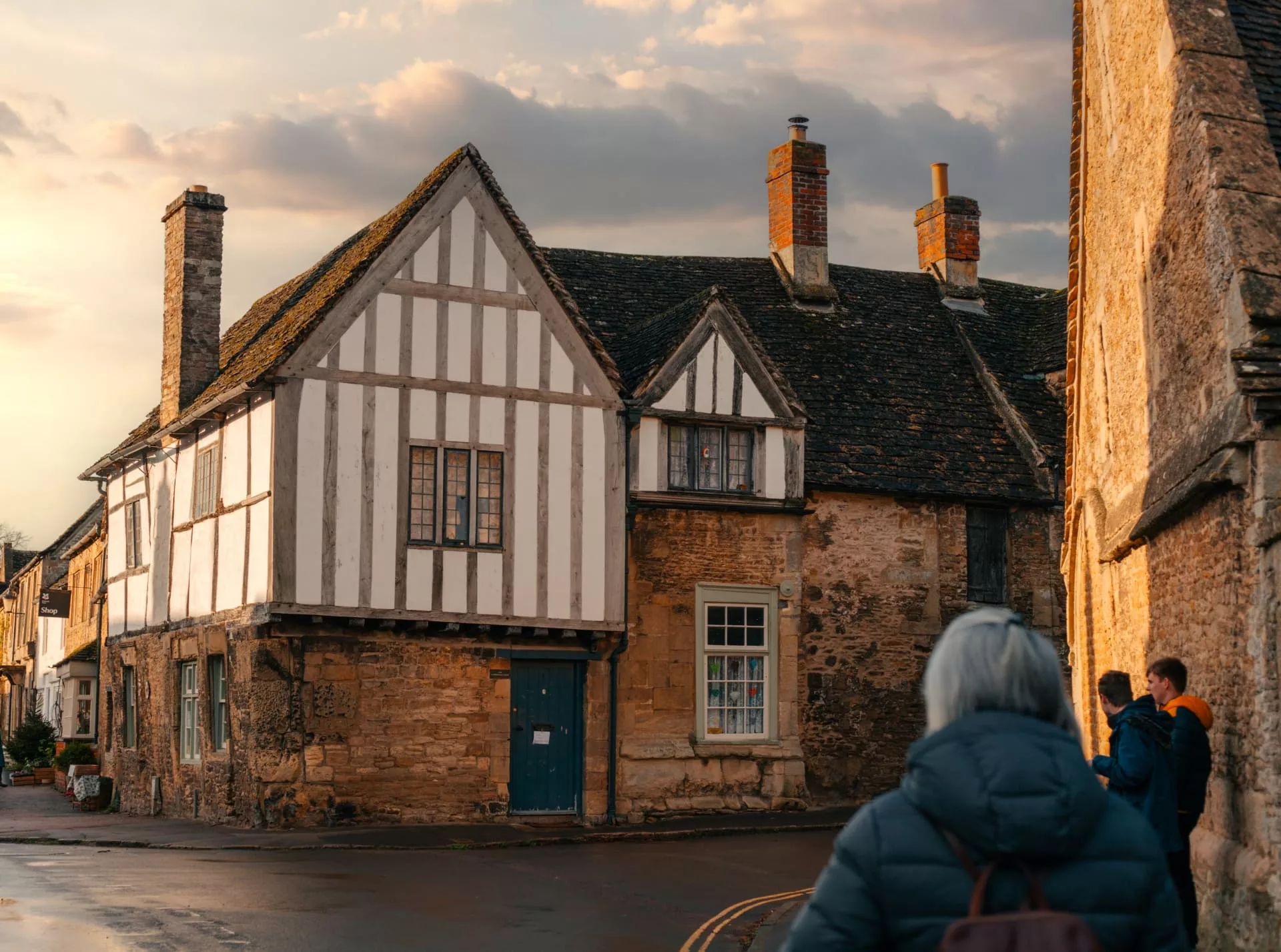 A street in Lacock with historic stone buildings. The sky is of pink hue and a woman and two young boys stand in the foreground of the image.
