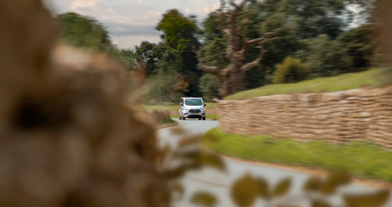 A van winds through a twisty lane where walls stand either side. There is foliage and trees in the foreground and background. Motion blur throughout the image.