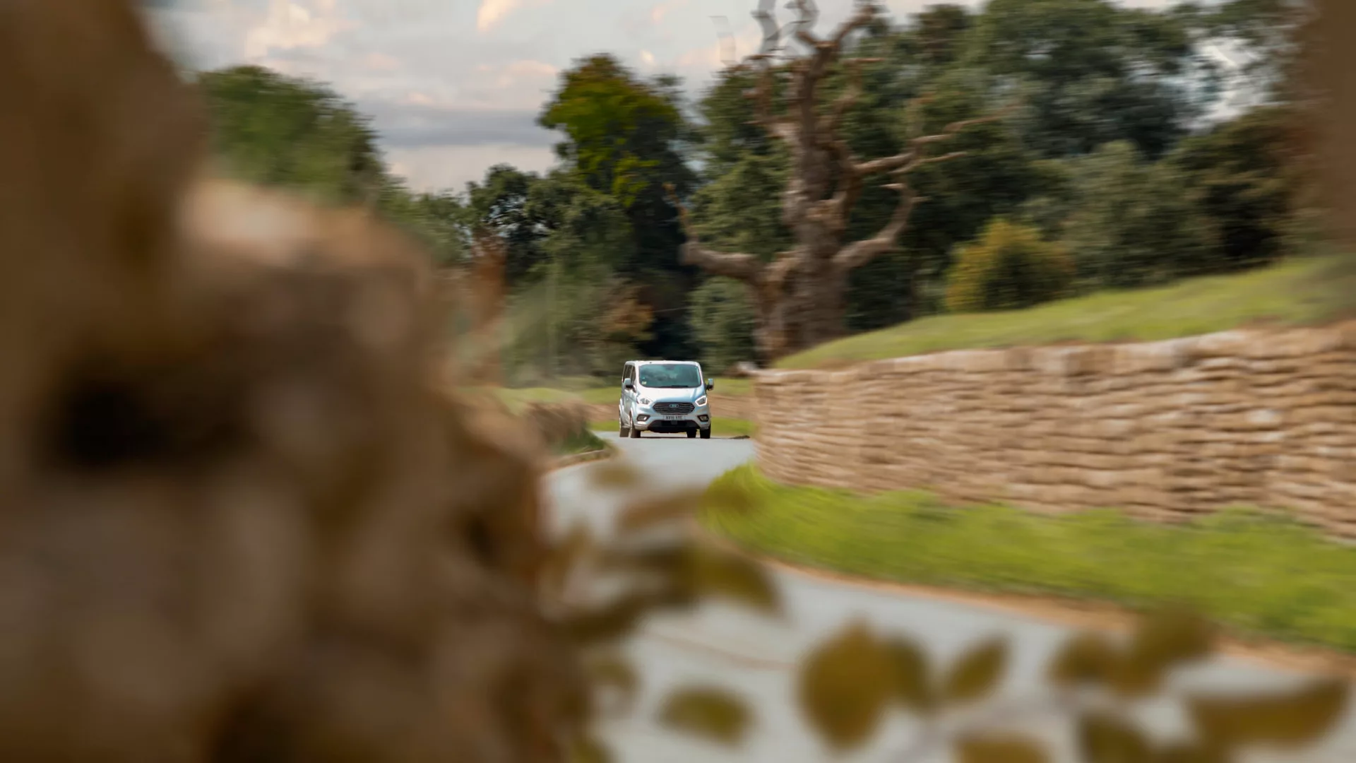 A van winds through a twisty lane where walls stand either side. There is foliage and trees in the foreground and background. Motion blur throughout the image.