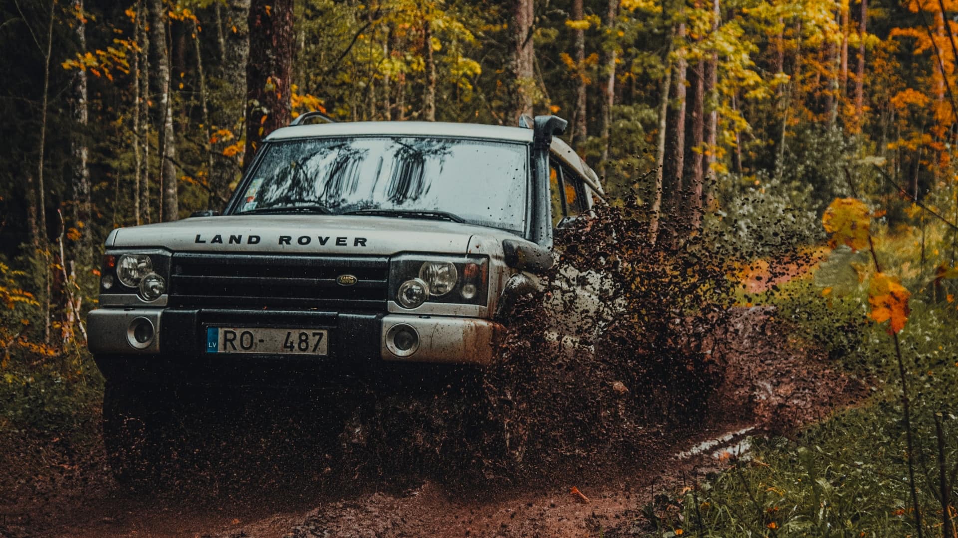 A Land rover plows through a muddy puddle. Surrounded by trees.