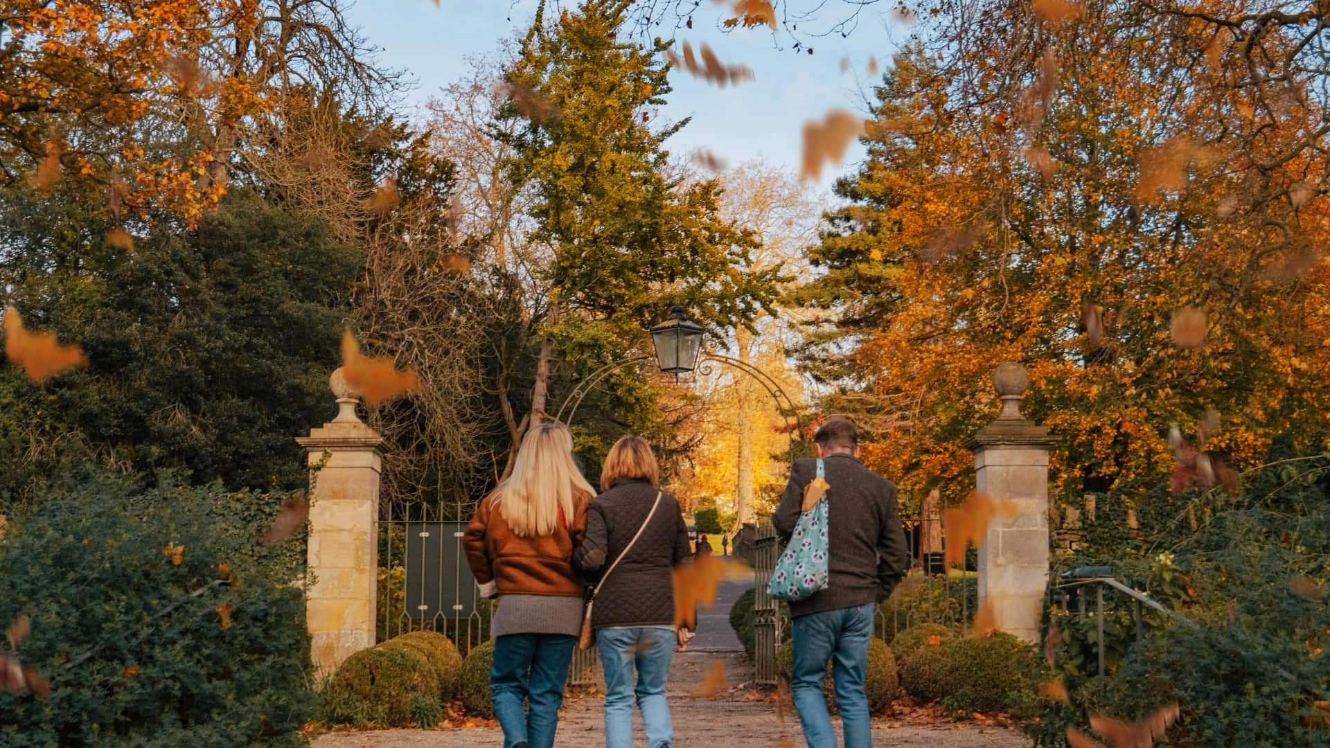 A beautiful autumn location with green, orange and yellow trees. A man, woman and daughter walk head on up the path with their backs to us.