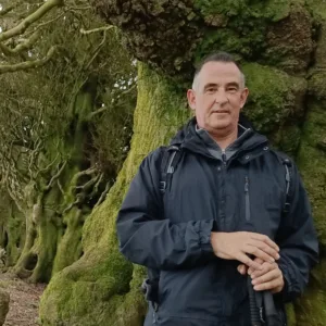 Man in a black jacket standing outdoors in front of a large moss-covered tree trunk.