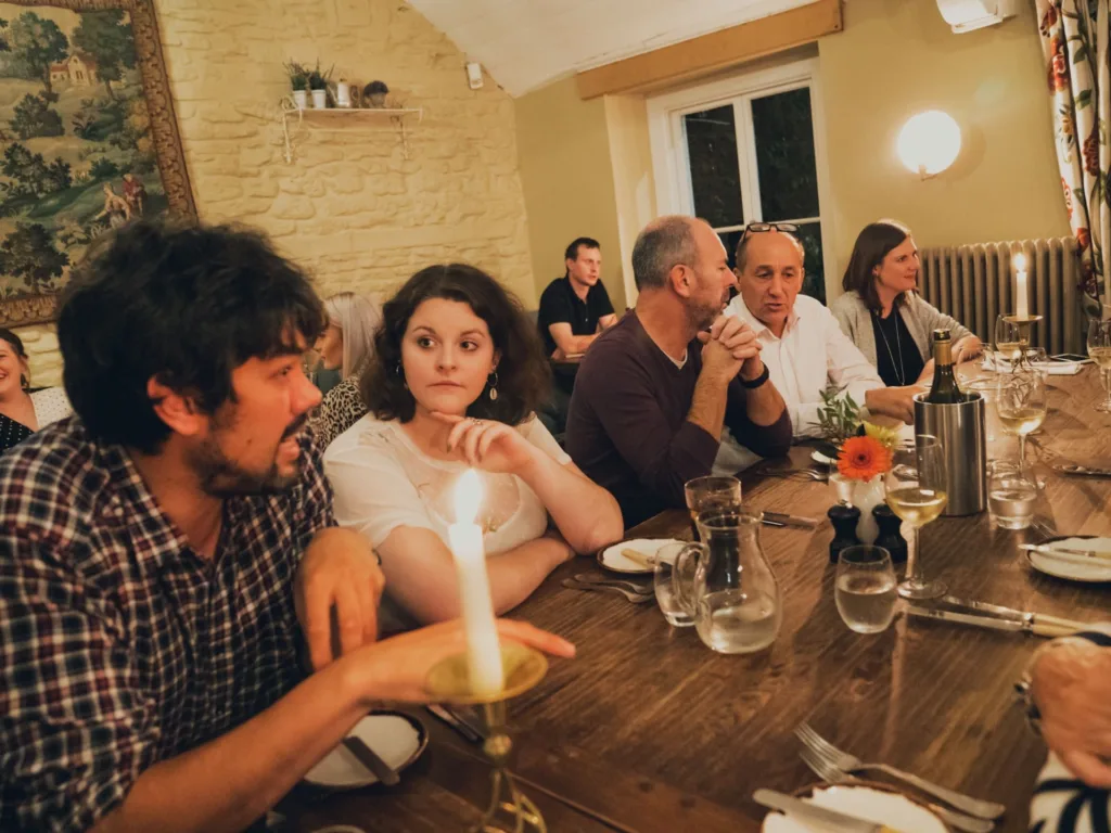 People sit round a long rectangular table. Different groups of discussions are happening and glasses of wine upon the table.