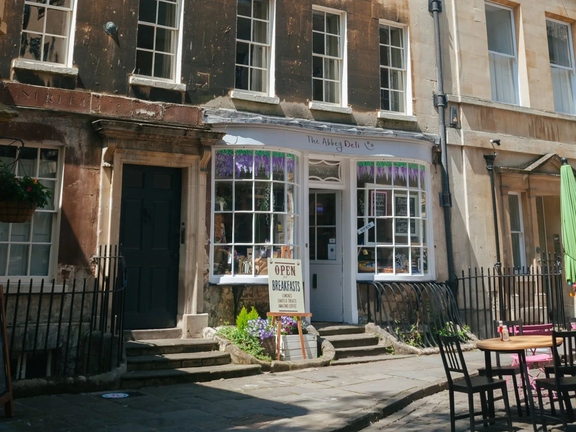 A photo of a small, quaint shop called "The Abbey Deli" in Bath, England. The building has a classic stone facade with large windows displaying various items inside. A small sign in front reads "OPEN BREAKFASTS." There are also outdoor seating areas with tables and chairs.