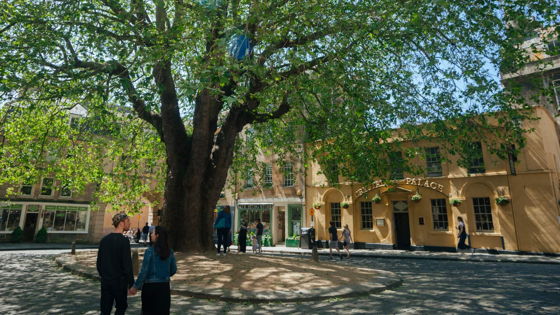 Couples walks hand-in-hand past the Crystal Palace pub on Abbey Green in Bath. A large, leafy tree provides shade as they stroll through the historic, Georgian architecture of the city centre.