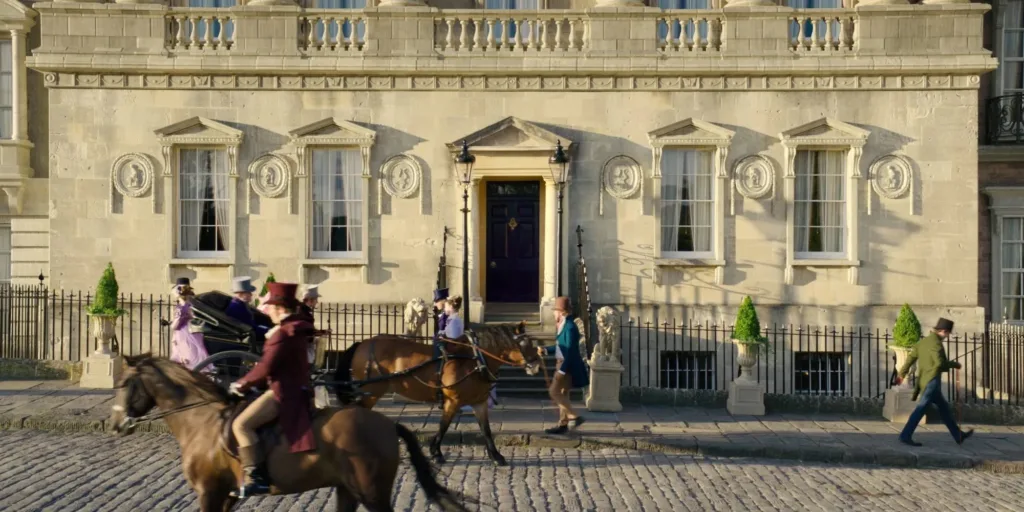 A scene from the Netflix series “Bridgerton” filmed at No.1 Royal Crescent in Bath, the filming location for the Featherington family home. The image shows actors in lavish Regency-era costumes walking along the iconic crescent.