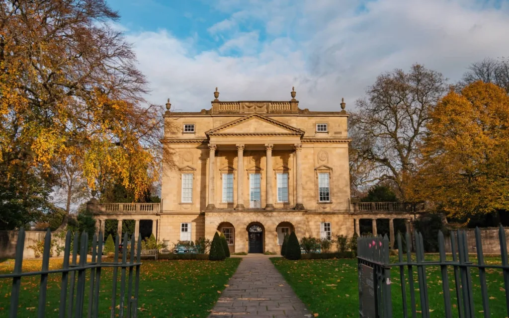 The Georgian façade of the Holburne Museum in Bath, framed by autumn trees and viewed from the garden path leading up to the entrance.