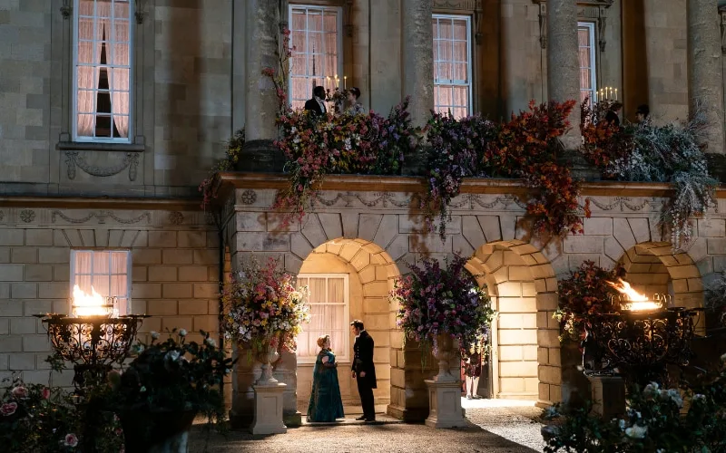 A scene from the Netflix series “Bridgerton” filmed at the Holburne Museum in Bath. The image shows the exterior of the museum, decorated with lavish floral arrangements, as Lady Danbury’s residence. Penelope Featherington and Colin Bridgerton can be seen interacting under an arch.