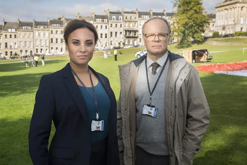 TV show "McDonald and Dodds" featuring Detectives McDonald and Dodds, played by Tala Gouveia and Jason Watkins, respectively. The duo stand in front of the iconic Royal Crescent in Bath.