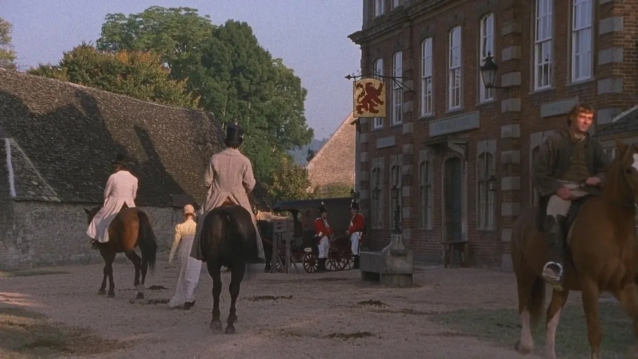 A view of a village setting where a large red brick building has a large, gold, lion sign suggesting it is an Inn. People line the street on horse back or walking along in period dress. This is Lacock village portrayed as Meryton in the 1995 Pride and Prejudice series.