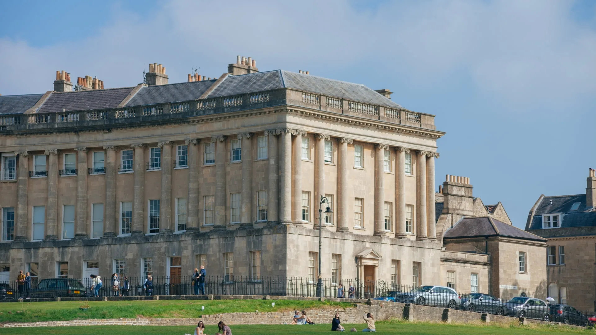 A view of the Royal Crescent in Bath, England. This iconic Georgian crescent of terraced houses, with their symmetrical design and elegant facades, is a popular tourist attraction. People can be seen walking along the street and relaxing on the surrounding lawns.