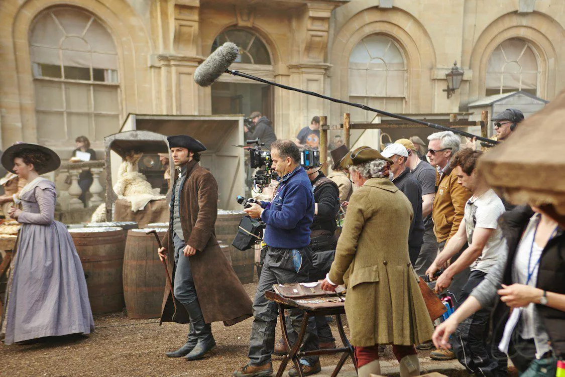 Aiden Turner as Captain Ross Poldark walking along a street in costume. A filming crew walks behind him. This street is in Corsham with cream stoned buildings in the background. The street is busy with extras.