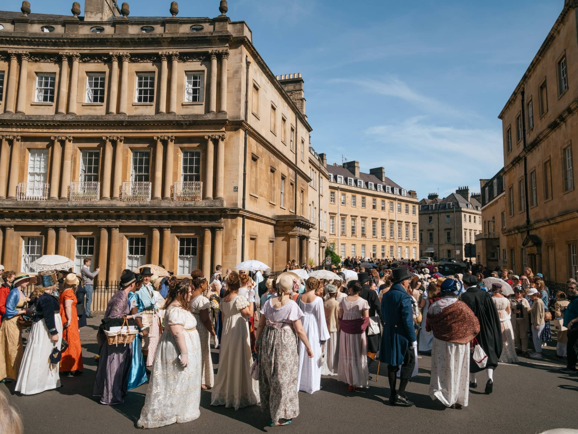 A bustling scene at the Circus in Bath, a circular Georgian plaza with grand, terraced townhouses. People are walking along the street and enjoying the vibrant atmosphere in period costume for the Jane Austen festival.