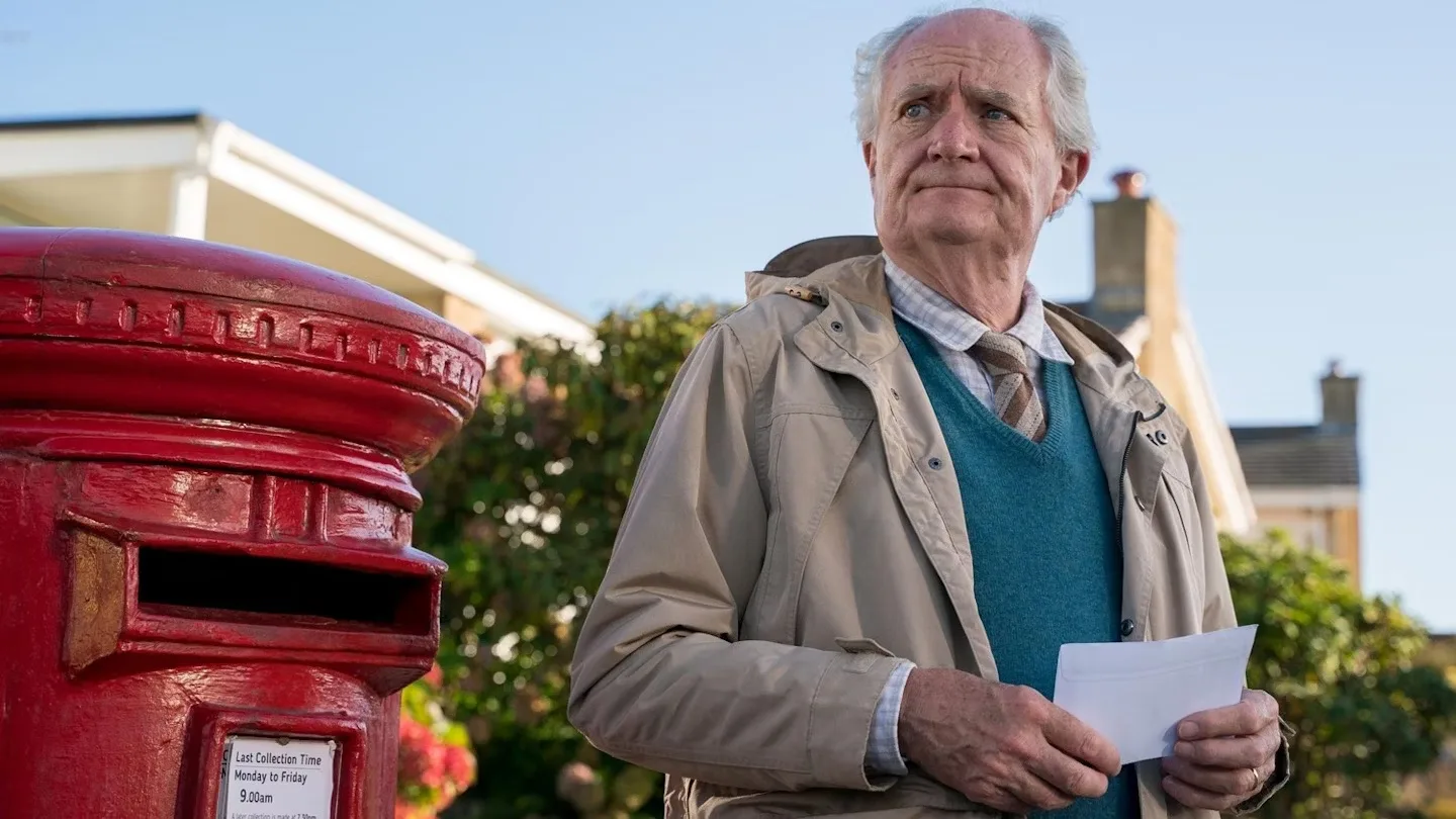 A scene from the film "The Unlikely Pilgrimage of Harold Fry" featuring Jim Broadbent as Harold Fry. The image shows Harold, an older man, standing next to a red British post box, looking contemplative.