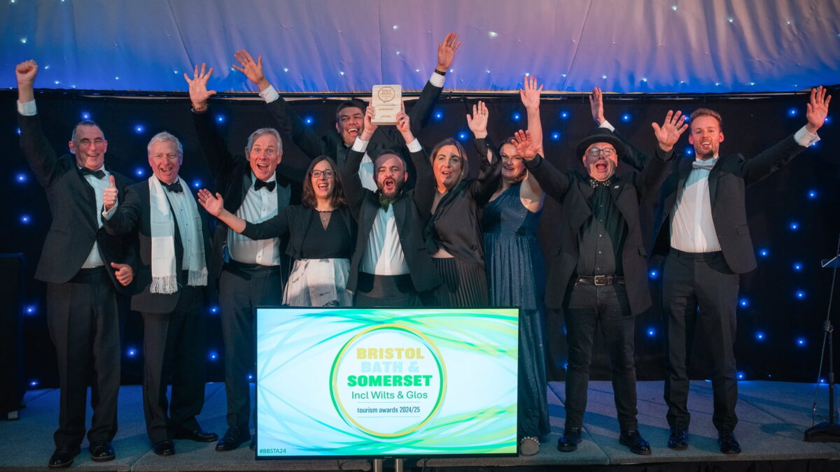 The team of In & Beyond Bath on the stage cheering. A sign in front of the stage reads 'Bristol, Bath & Somerset including Wilts & Glos Tourism Awards 2024/25'.