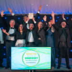 The team of In & Beyond Bath on the stage cheering. A sign in front of the stage reads 'Bristol, Bath & Somerset including Wilts & Glos Tourism Awards 2024/25'.