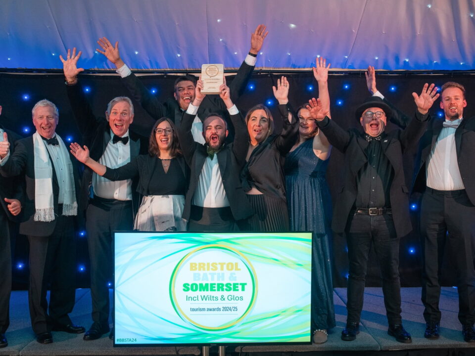 The team of In & Beyond Bath on the stage cheering. A sign in front of the stage reads 'Bristol, Bath & Somerset including Wilts & Glos Tourism Awards 2024/25'.