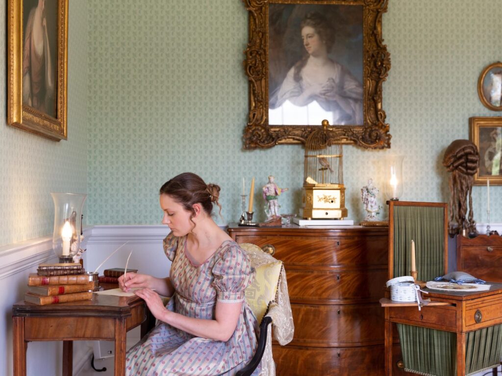 Woman in Regency-era dress sitting at a small wooden desk, writing with a quill by candlelight in a period-decorated room with antique furniture and portraits.