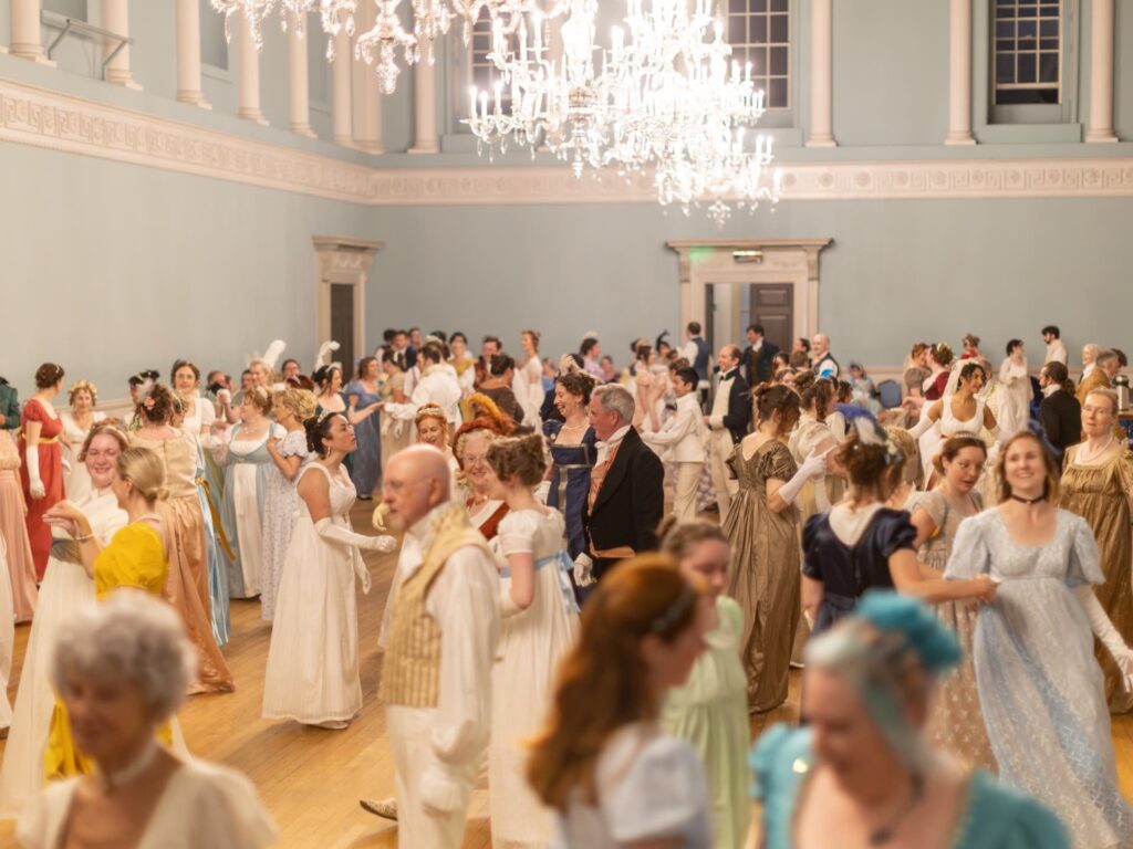 Large ballroom filled with people in Regency-era costumes dancing beneath chandeliers during a Jane Austen-themed event.